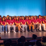 A large group of students in matching maroon T-shirts sit on stage in rows, facing an audience. The background is bright blue, and some audience members are visible in the foreground.