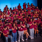 A large group of students wearing matching maroon t-shirts sit and stand on a stage, clapping and smiling in front of a blue backdrop. Some students are standing while others are seated in rows.