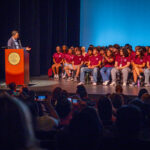 A man speaks at a podium on a stage while a group of students in matching maroon shirts sit and listen; an audience in the foreground watches and takes photos.