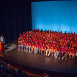 A large group of students in matching maroon shirts sit onstage in rows, facing an audience. A person stands at a podium on the left, speaking, with a blue-lit background behind the students.