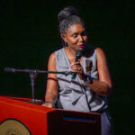 A woman stands at a podium, holding a microphone and speaking. She wears a sleeveless gray top and has her hair in braids tied up. She appears engaged with her audience, illuminated against a dark background.