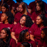 A group of young people wearing matching maroon shirts sit closely together in rows, some looking around and others talking or lost in thought, against a blue background.