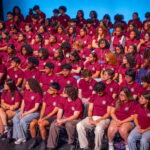 A large group of young people wearing matching maroon t-shirts sit and stand together on tiered risers against a blue background, posing for a group photo. Most are facing forward, some are looking at each other.