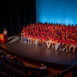 A large group of people in matching maroon shirts sits on stage in front of a blue background, while a speaker stands at a podium addressing an audience in a theater.
