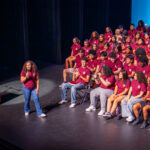 A woman speaks on stage with a microphone in front of a large group of students wearing matching maroon shirts, seated and smiling. A podium stands to the side, and the background is lit in blue.