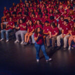A young woman stands center stage with a microphone, addressing a large group of students seated behind her. All the students wear matching maroon shirts and jeans or khakis, suggesting a school or team event.