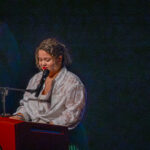 A woman with curly hair, wearing a patterned white blouse, stands at a podium with a microphone. The podium has a circular gold emblem. The background is dark, and she appears to be speaking or reading.