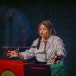 A woman with curly hair, wearing a white blouse and green skirt, speaks passionately at a podium with a microphone, gesturing with her hands against a dark background.