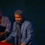 A man in a dark suit speaks into a microphone at a podium, while a woman in a white blouse stands in the background holding a folder, watching him. The background is dark and the scene is brightly lit.
