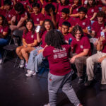 A group of students in matching maroon shirts sit in rows of chairs, some holding up phones and smiling as a person in the foreground, also in maroon, faces them and gestures enthusiastically.