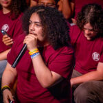 A person with curly hair speaks into a microphone, wearing a maroon EOF Achieve shirt and colorful wristbands, surrounded by others in matching shirts seated nearby.