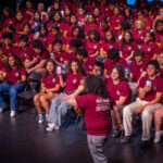 A large group of students in matching maroon t-shirts sits in rows on a stage, holding up lit cell phones. One person stands facing the group, speaking or performing. The atmosphere appears lively and engaged.