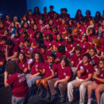 A large group of students in matching maroon T-shirts sit on stage, many holding up lit cell phones. The background fades from blue to black, and a person stands in front facing the group.