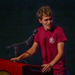 A young man in a maroon t-shirt stands at a red podium, speaking into a microphone, against a dark background. He is looking to the side while holding the podium with one hand.