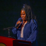 A woman with long braided hair stands at a podium, speaking into a microphone. She is wearing a blue pinstripe jacket and is illuminated against a dark background.