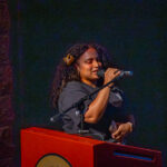 A woman with curly hair, wearing a dark shirt and a flower hair clip, speaks into a microphone while standing at a red podium against a dark background.