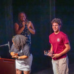 A young man smiles and holds a glass award on stage while a woman claps and another woman stands at a podium, adjusting a microphone. The setting appears to be a college event or award ceremony.