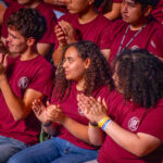 A group of young people wearing matching maroon shirts sits closely together, clapping and smiling, appearing engaged and supportive at an event.