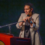 A woman in a beige blazer speaks into a microphone at a podium during a presentation or event, standing against a dark background.