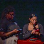 Two women stand at a podium; one holds a phone and microphone while appearing emotional, the other looks at a tablet. Both are focused on their devices against a dark background.