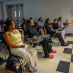 A woman stands and speaks in front of a classroom of students seated at desks, listening attentively. The classroom has posters on the walls and natural light coming through the windows.