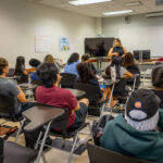 A classroom with students seated at desks, facing two women standing at the front. One woman is speaking while the other stands nearby. Posters are on the walls and a large monitor is beside them.