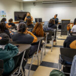 A classroom full of students seated at desks, facing two instructors at the front. One instructor is standing and speaking, while the other is seated. The students appear attentive, and a whiteboard is visible in the background.