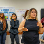 A woman speaks in front of a group of four students standing by a wall with posters and charts in a classroom. The students listen attentively, some with their arms crossed or hands near their faces.