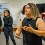 A woman with curly hair speaks while gesturing with her hands in a classroom. Four students stand in the background near posters and a computer, listening attentively.