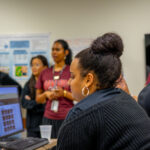 Five women are in a classroom setting. One is seated at a computer, focused on the screen, while four others stand nearby, talking and looking engaged. Posters are visible on the wall in the background.