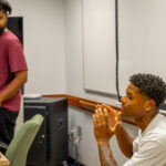 Two men are in a classroom; one in a maroon shirt stands near a desk, while another in a white shirt sits, speaking with his hands raised. A notebook and green soda bottle are on the table.