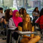 A diverse group of students sits attentively at desks in a bright classroom, with notebooks and pens on their desks. Sunlight streams through large windows in the background.