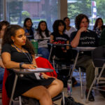 A diverse group of students sits attentively in a classroom, some taking notes and others listening. Desks are arranged in rows, and sunlight shines through large windows in the background.