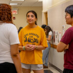 Three students stand in a hallway, talking. One wears a yellow Levi Strauss t-shirt and shorts, another wears a white shirt, and the third wears a maroon shirt and glasses. Bulletin boards and posters are visible on the walls.
