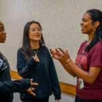 Three young women stand in a hallway, smiling and talking to each other. Two women wear black sweatshirts, and one wears a maroon t-shirt. They appear to be engaged in a friendly conversation.