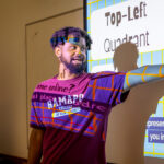 A man in a maroon Ramapo College t-shirt stands in front of a projected presentation, pointing to the screen with Top-Left Quadrant displayed. The projections grid and text are cast over his face and shirt.