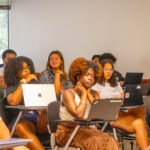 A group of students sit in a classroom, listening attentively to a speaker. Several have laptops open on their desks, and large windows show greenery outside. The atmosphere appears focused and engaged.