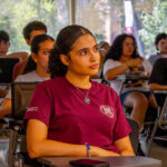 A young woman in a maroon shirt sits attentively at a desk in a classroom, surrounded by other students with laptops. Sunlight streams through large windows behind them.