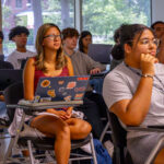 A group of students sit in a classroom, listening attentively. Several have laptops open, and one student in the front has a laptop covered in colorful stickers. Large windows show trees and greenery outside.