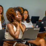 A group of students sit in a classroom, listening attentively. Several have laptops open on their desks. The focus is on a student in the foreground with curly hair, resting her hands on her neck and looking thoughtful.