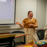 A woman stands and smiles while giving a presentation in a classroom. She gestures with one hand toward a projection screen. Nearby are desks, a laptop, and a bowl of colorful pens.