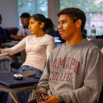 A group of college students sit at desks in a classroom, listening attentively. Some take notes, while others watch the front of the room. Laptops and notebooks are on the desks, and large windows provide natural light.