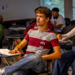 A group of students sit at desks in a classroom, attentively facing forward. Some have notebooks, drinks, and pens, and large windows let in natural light. The mood appears focused and engaged.