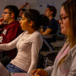 Students sit attentively at desks in a classroom, focusing on something off-camera. Some take notes or have laptops open, while warm lighting highlights their engaged expressions. Posters and writing are visible on the back wall.