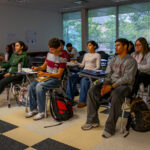 A group of students sit at desks in a classroom, attentively facing forward. Notebooks and backpacks are on their desks and the floor. Large windows in the background let in natural light.
