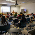 A classroom with students seated at desks facing a teacher presenting slides at the front. The teacher gestures toward the projected screen while students listen and take notes. The room has white walls and fluorescent lighting.