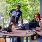 Five students are gathered around a table outdoors, using laptops and books, studying or working together. Trees and greenery are visible in the background.