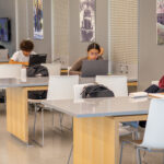 Several students sit at tables in a modern study area, working on laptops and tablets. Backpacks are on the tables, and historical photos hang on the gray walls in the background.