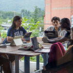 Five students sit around a table outdoors, talking and working on tablets and laptops, with greenery and a building visible in the background. The group appears engaged in discussion.