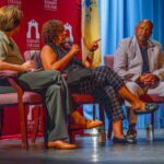Three people sit on a stage in front of a red Ramapo College backdrop, engaging in conversation. The woman in the center gestures while speaking, as the other two listen attentively. A flag is visible in the background.